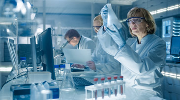 Scientists in protective lab gear analyzing samples and data in a modern laboratory environment, with test tubes and computers visible on the desks.