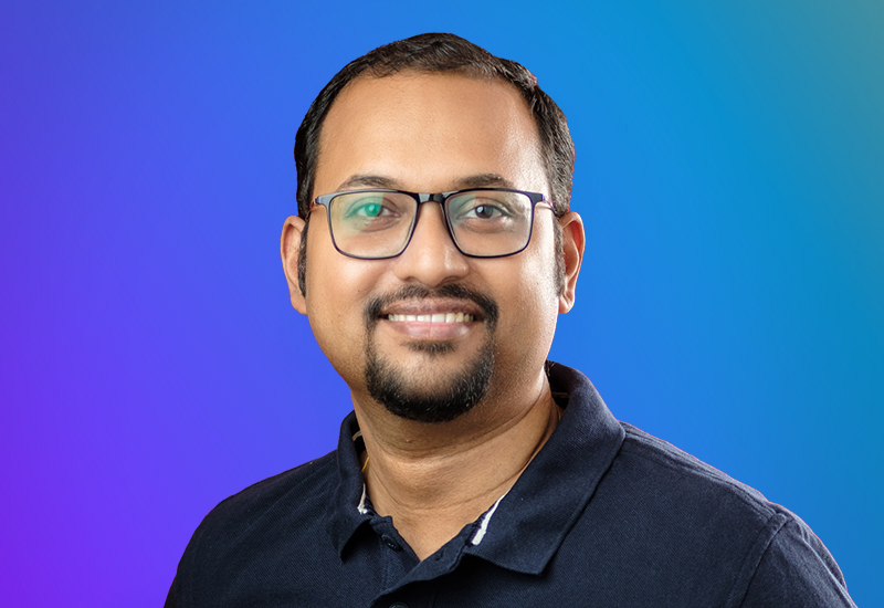 Portrait of a smiling man wearing glasses and a dark collared shirt, with a gradient blue background.