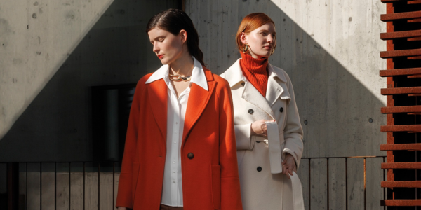 Two individuals wearing stylish coats, one in bright orange and the other in cream, standing in a modern architectural setting with dramatic sunlight and shadows.