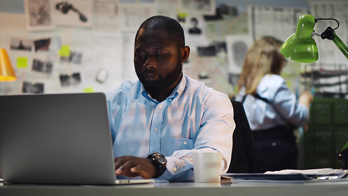 African detective processing evidence working on laptop in office. Portrait of serious afro-american police office sitting at desk and using computer at police station
