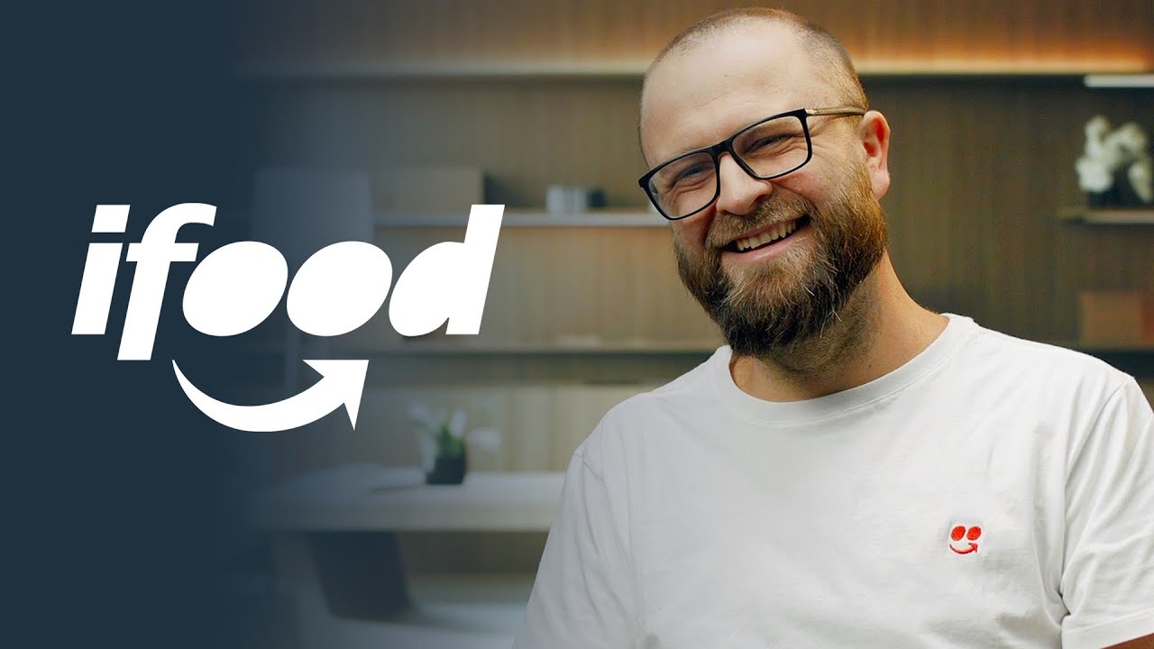 A bearded man with glasses smiling, wearing a white shirt with a small logo, is seated indoors with a blurred background. The left side features the prominent iFood logo in white on a dark overlay.