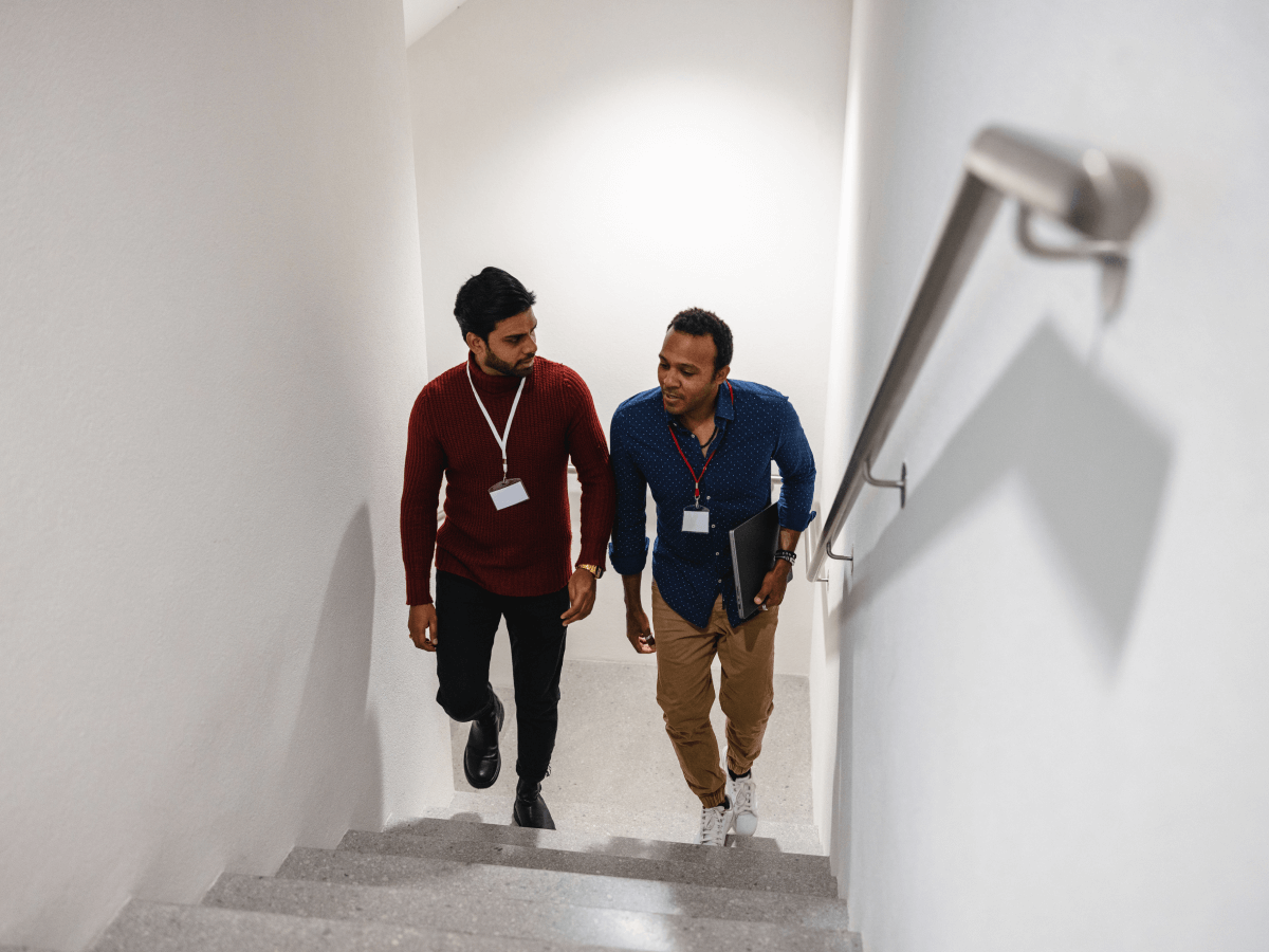 Two men wearing name badges are walking up a staircase in a modern indoor office setting. One holds a folder, and both appear to be engaged in conversation.
