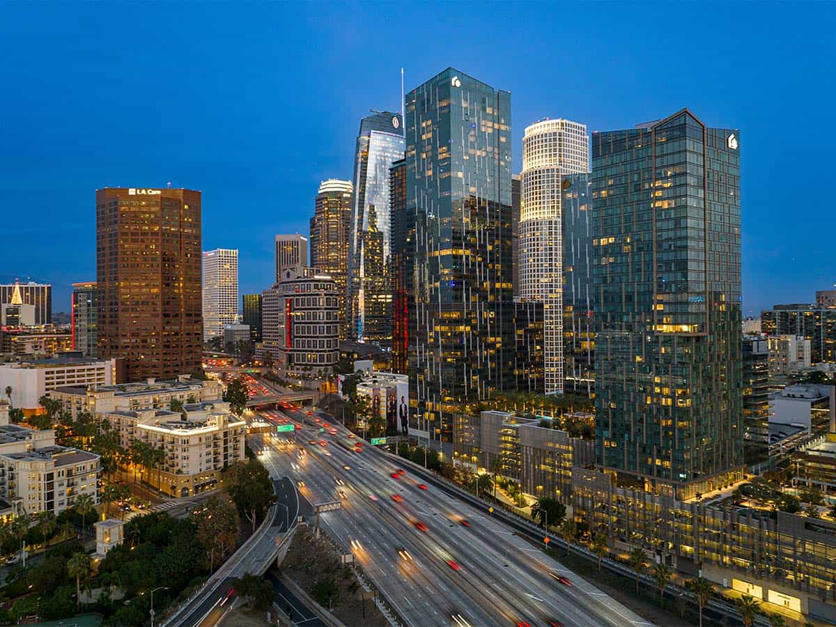 A vibrant modern city skyline featuring tall glass skyscrapers and a busy highway at dusk, with building lights and vehicle motion creating a dynamic urban atmosphere.