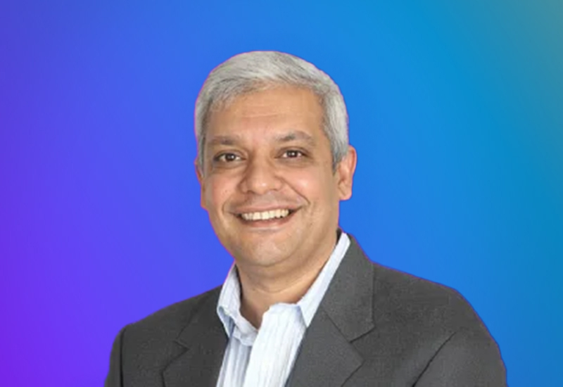A portrait of a smiling man with gray hair, wearing a suit and light-colored shirt, set against a blue gradient background.