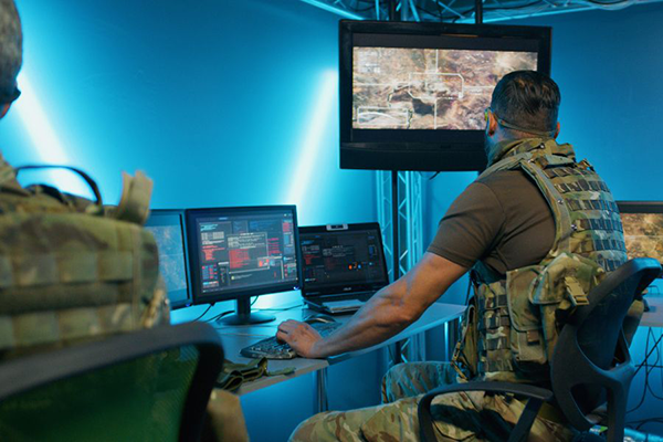 Two military personnel in camouflage uniforms monitor surveillance data and battlefield activity on multiple computer screens and a large wall monitor in a control room with blue lighting.