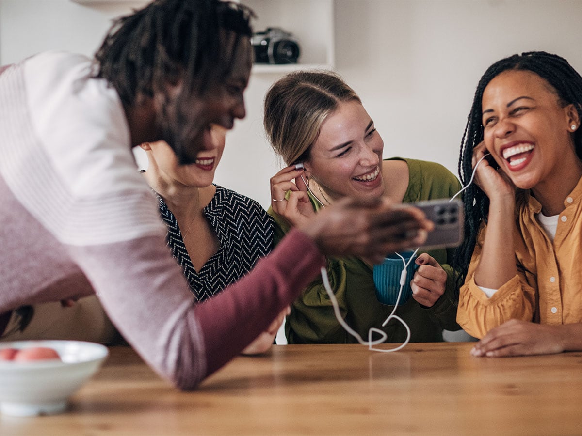 A group of women sitting at a table, sharing earphones and laughing together while listening to music on a smartphone.