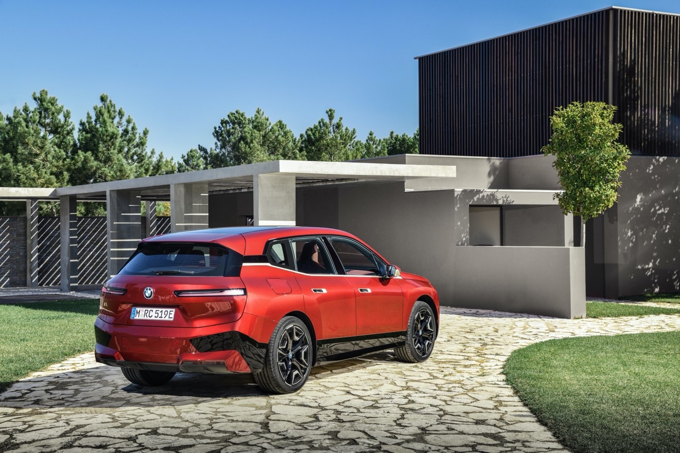 A red SUV is parked on a stone driveway in front of a modern home with geometric architecture, surrounded by greenery and trees under a clear blue sky.