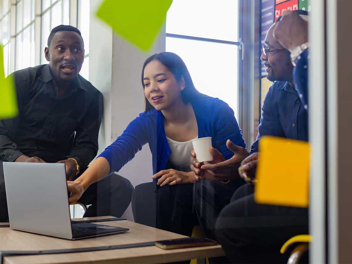 Three colleagues collaborating around a laptop in a modern office space, discussing work and sharing ideas.