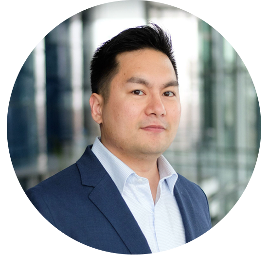 A business professional male in a blue suit and white shirt, posing for a headshot in a modern office environment with glass walls in the background.