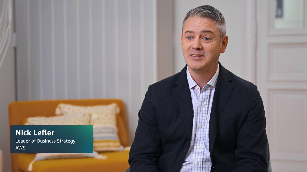YouTube thumbnail image for an AWS business strategy video, showing a professionally dressed individual seated indoors with an orange couch and pillows in the background. The left corner overlay displays 'Nick Lefler, Leader of Business Strategy, AWS.'