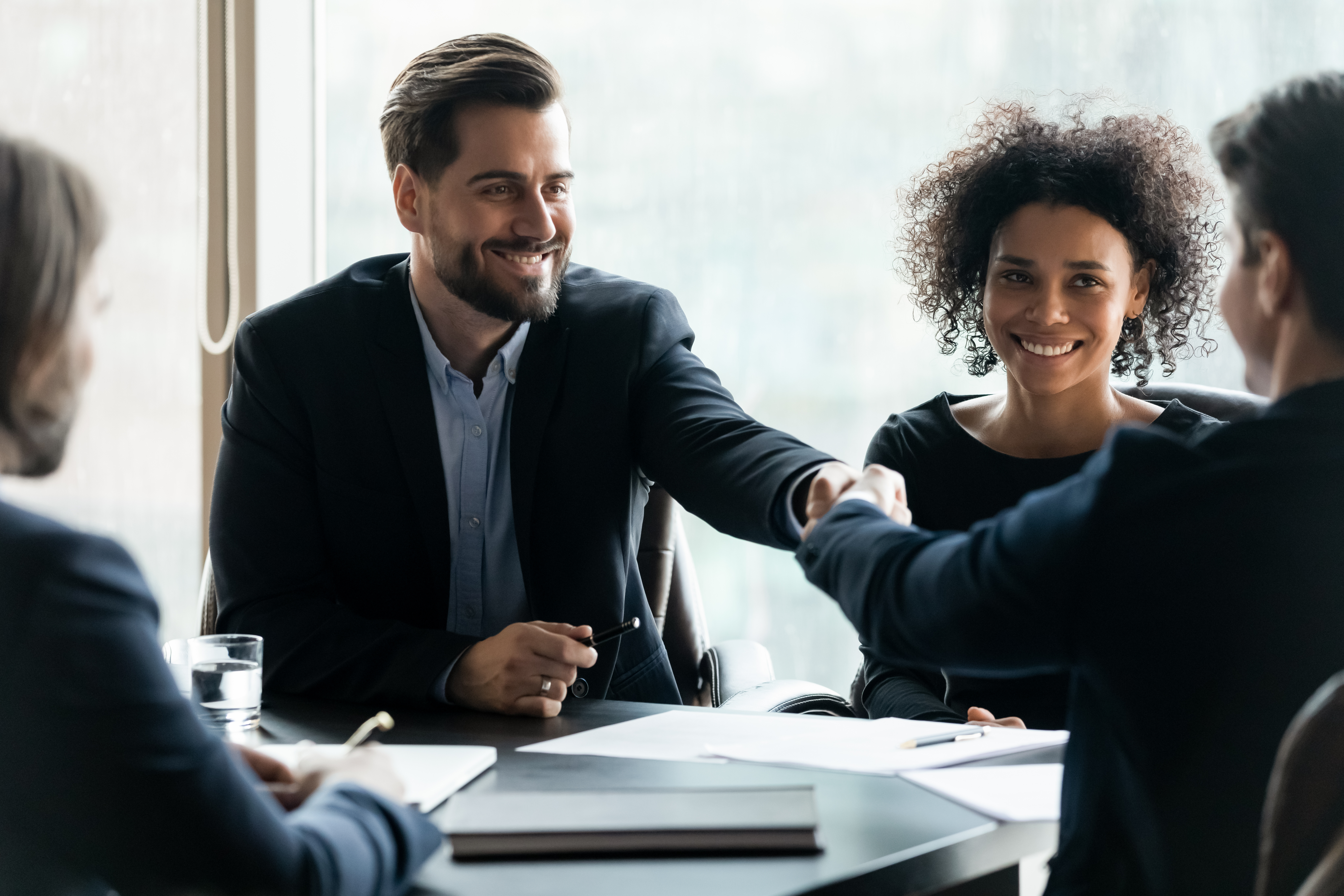 Business professionals sitting around a table in a modern office, with two people smiling and shaking hands to celebrate a successful partnership or agreement.