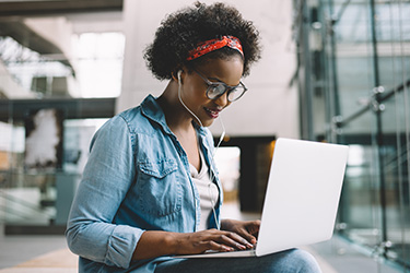 A woman wearing glasses and headphones is smiling while working on a laptop in a modern office or workspace environment.