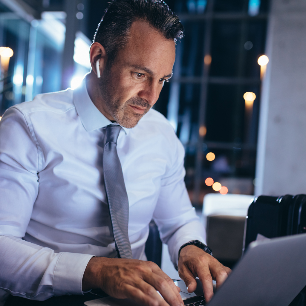 Man in a white shirt and tie typing on a laptop in a modern, dimly lit office setting.