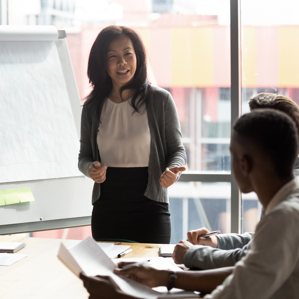 A woman is smiling and presenting during a business meeting, standing near a flip chart while colleagues listen and take notes.