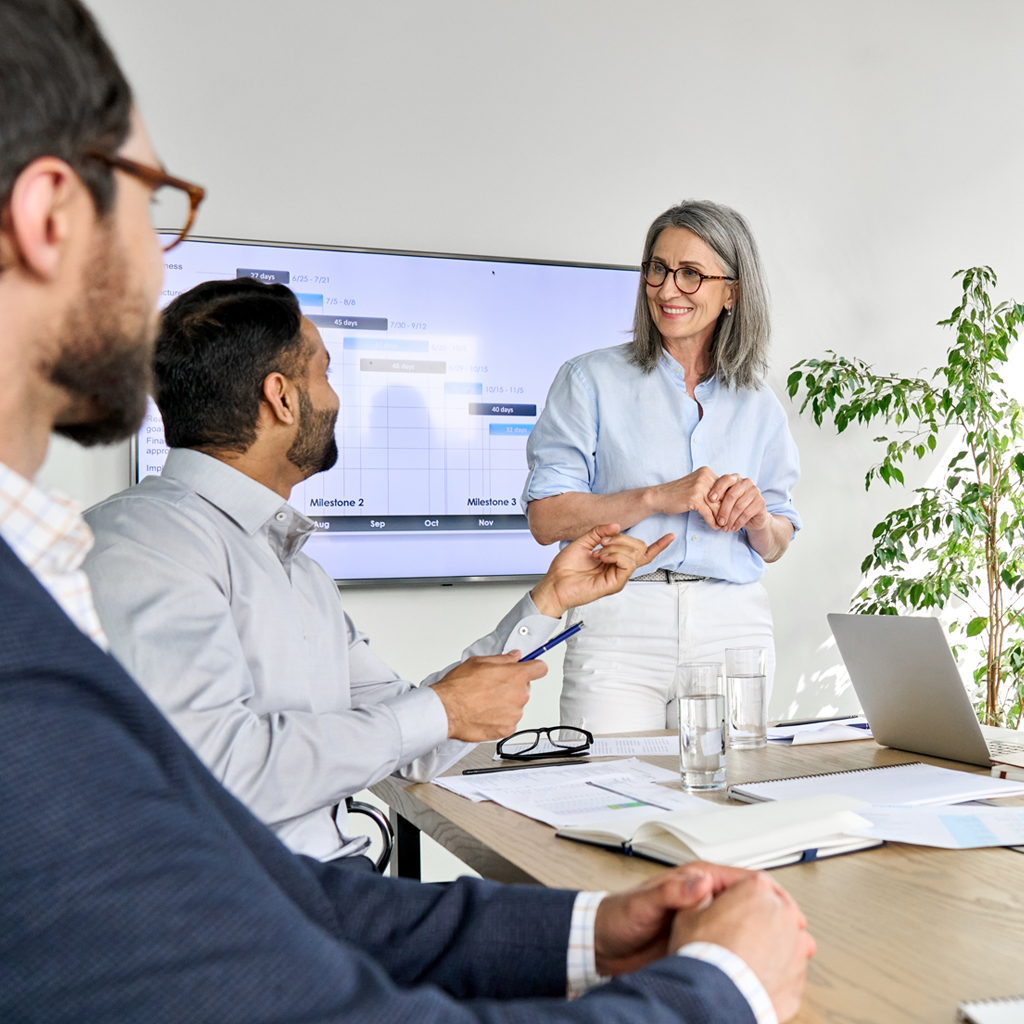 A woman smiling and leading a business meeting presentation with colleagues. A screen with a project timeline is visible in the background, and business documents are on the table.