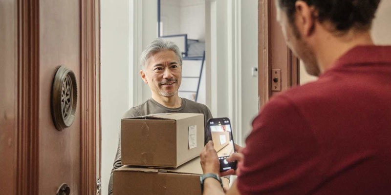 A man receives two cardboard packages at his home door while another person, possibly a delivery worker, takes a photo of the delivery with a mobile phone.