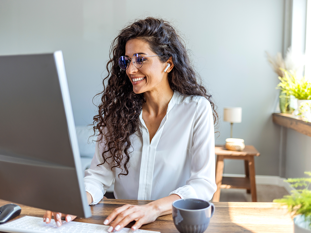 A businesswoman wearing glasses and earbuds is smiling while working on a computer in a modern office setting with natural light.