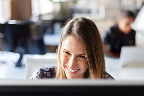 A woman smiling while working on a computer in a modern office environment.