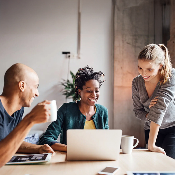 Three colleagues collaborating around a table with a laptop and coffee mugs, smiling and discussing together in a modern office environment.