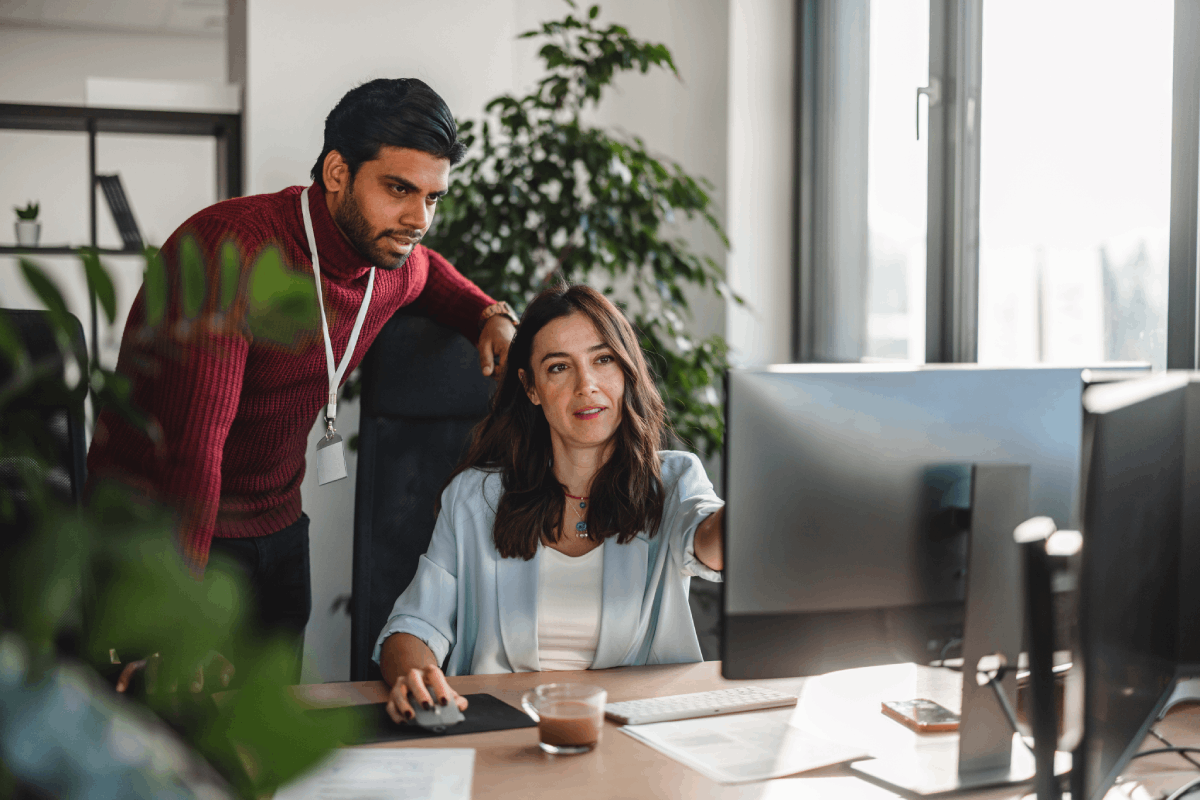 Two coworkers collaborating at a computer workstation in a modern office environment with large windows and indoor plants.