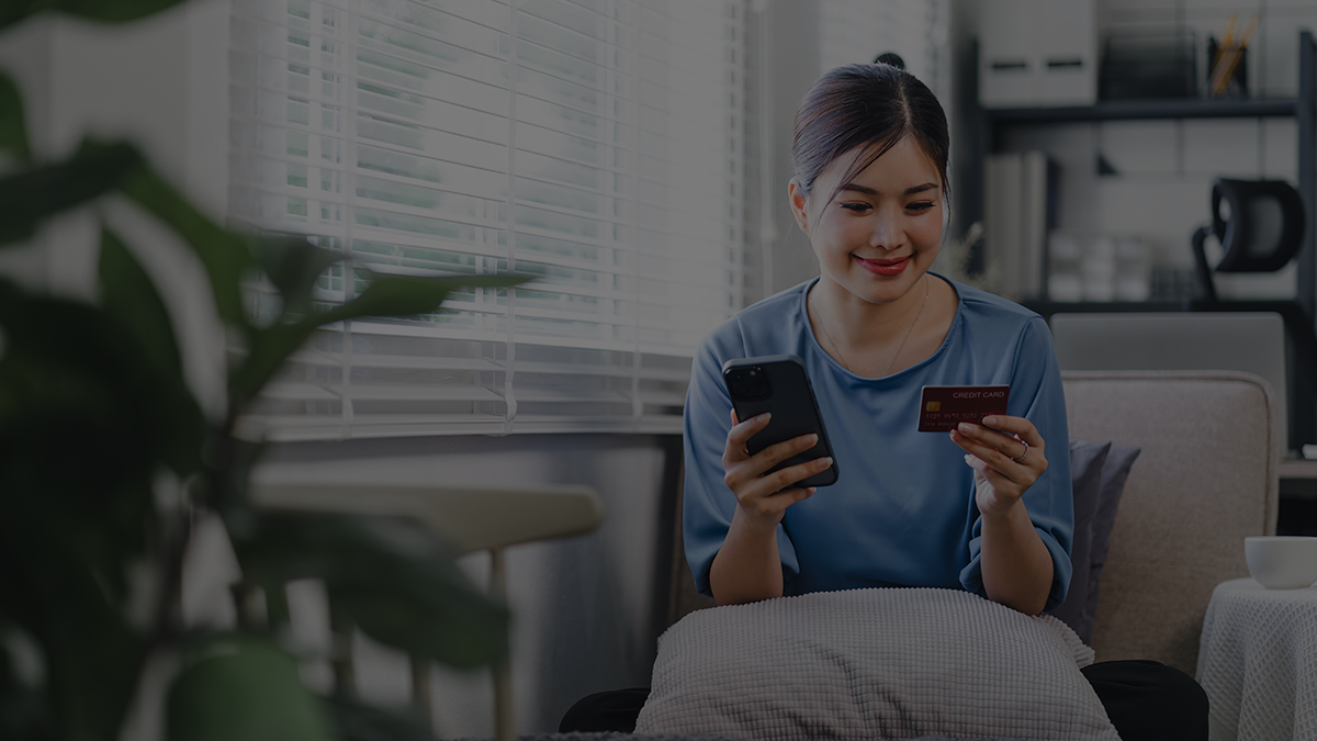 A woman sitting in a living room, holding a credit card and a smartphone, making an online purchase. She is smiling and appears relaxed, with home furnishings in the background.