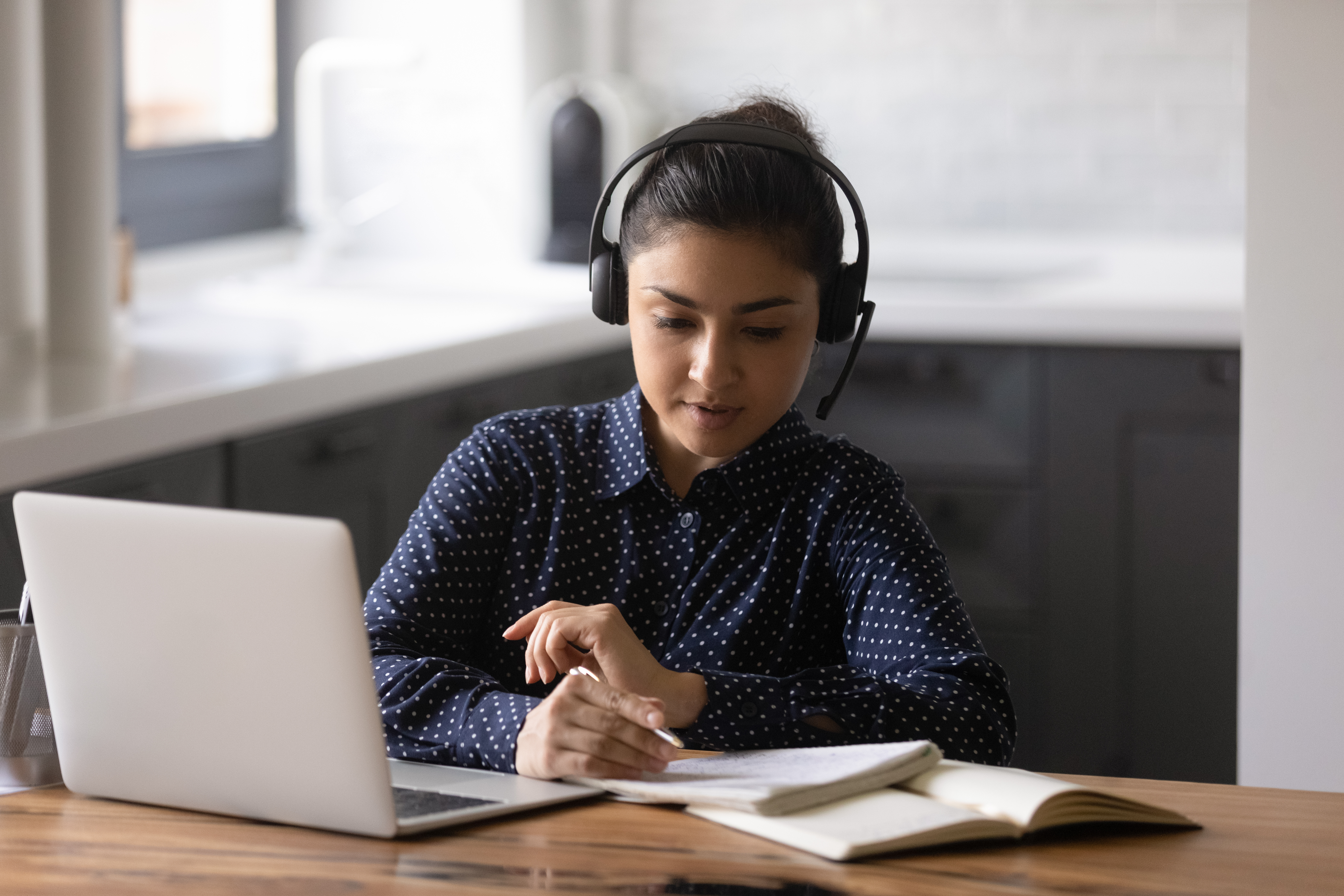 E-learning. Concentrated indian female student sitting by pc at home office wearing headphones. Young hindu woman listening to video lecture writing notes to copybook by hand. Getting education online