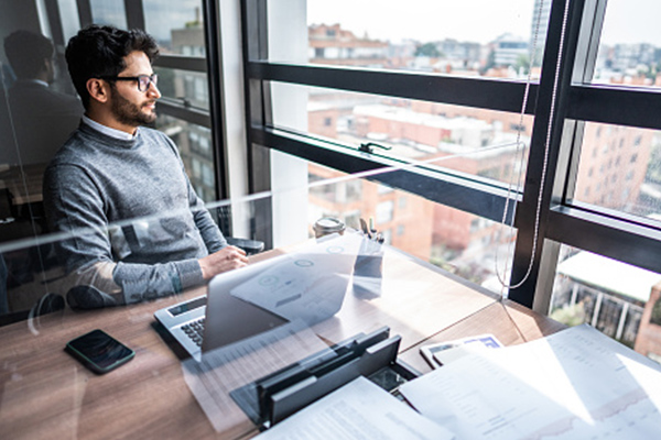 A businessman sitting at a desk in a modern office, looking out the window with a city view, surrounded by documents and a laptop.