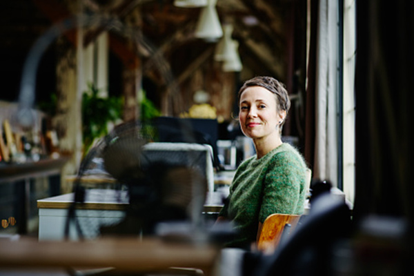 A woman in a green sweater sits at a desk in a modern, well-lit office with exposed beams and large windows.