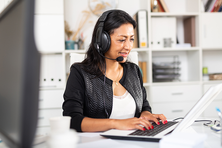A woman wearing a headset is working at her computer in a modern office environment, possibly providing customer service or support.