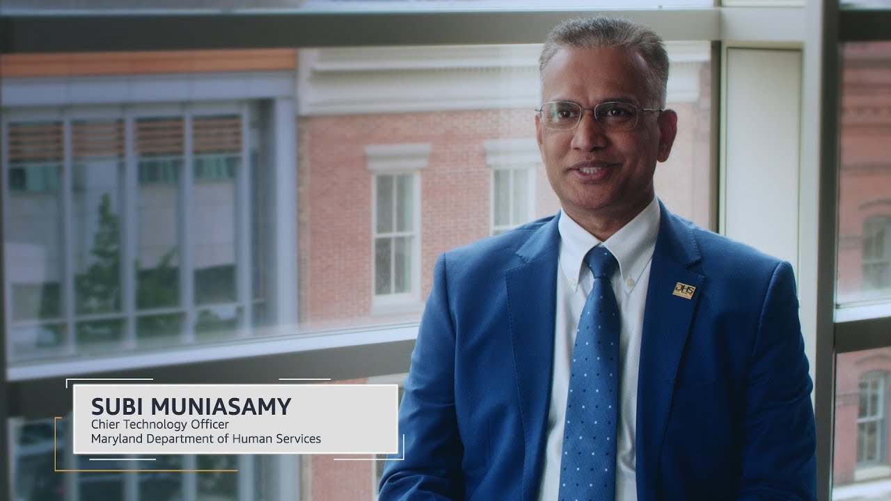 A person in a blue suit and tie sitting indoors near a window, with text identifying Subi Muniasamy as Chief Technology Officer, Maryland Department of Human Services.
