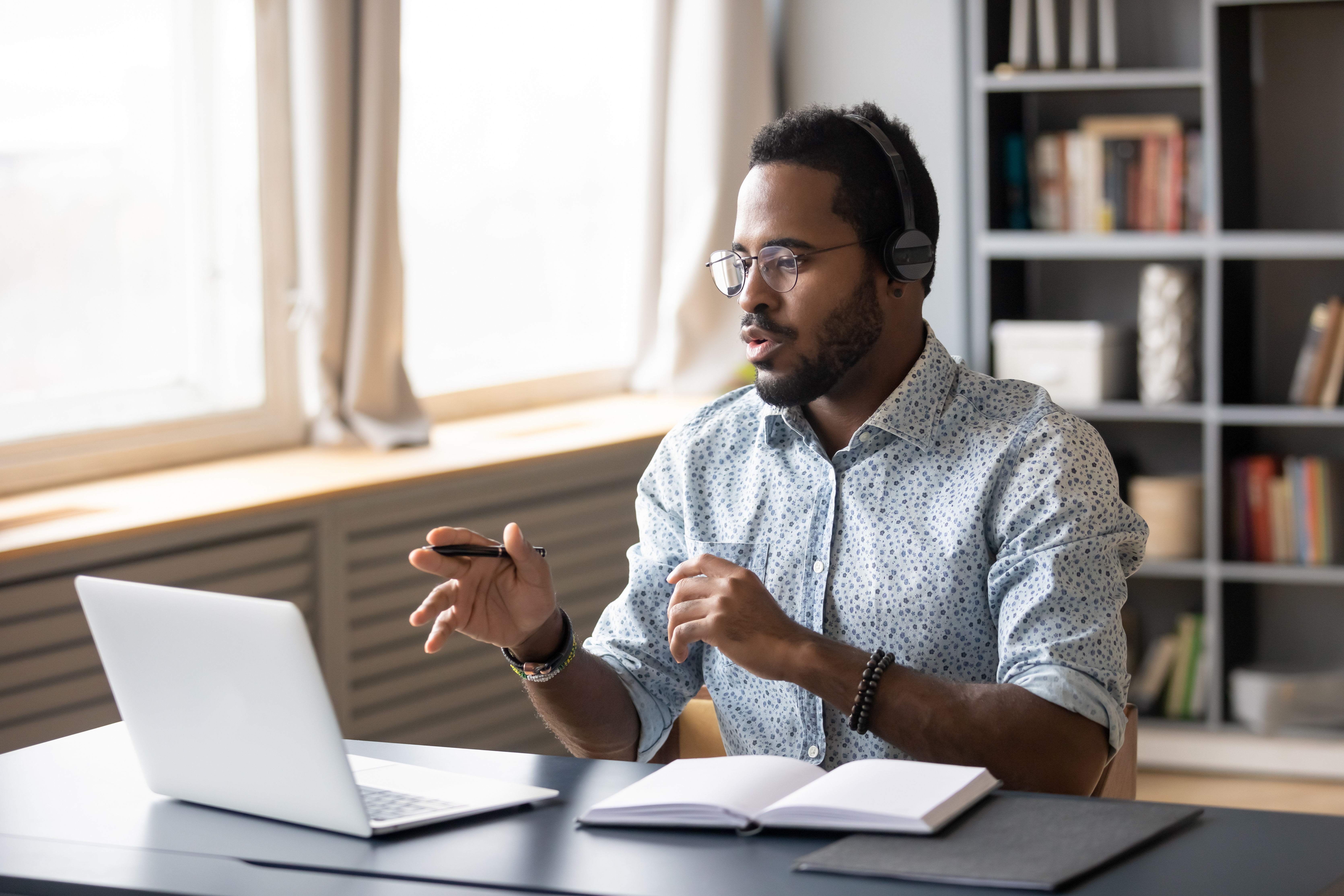 Person wearing headphones gesturing with a pen during a video call, seated at a desk with a laptop and open notebook.
