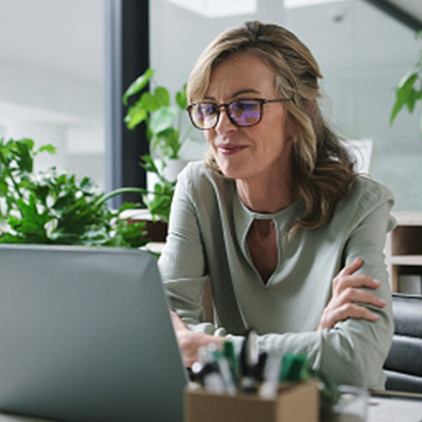 A woman wearing glasses works on a laptop in a modern office surrounded by green plants.