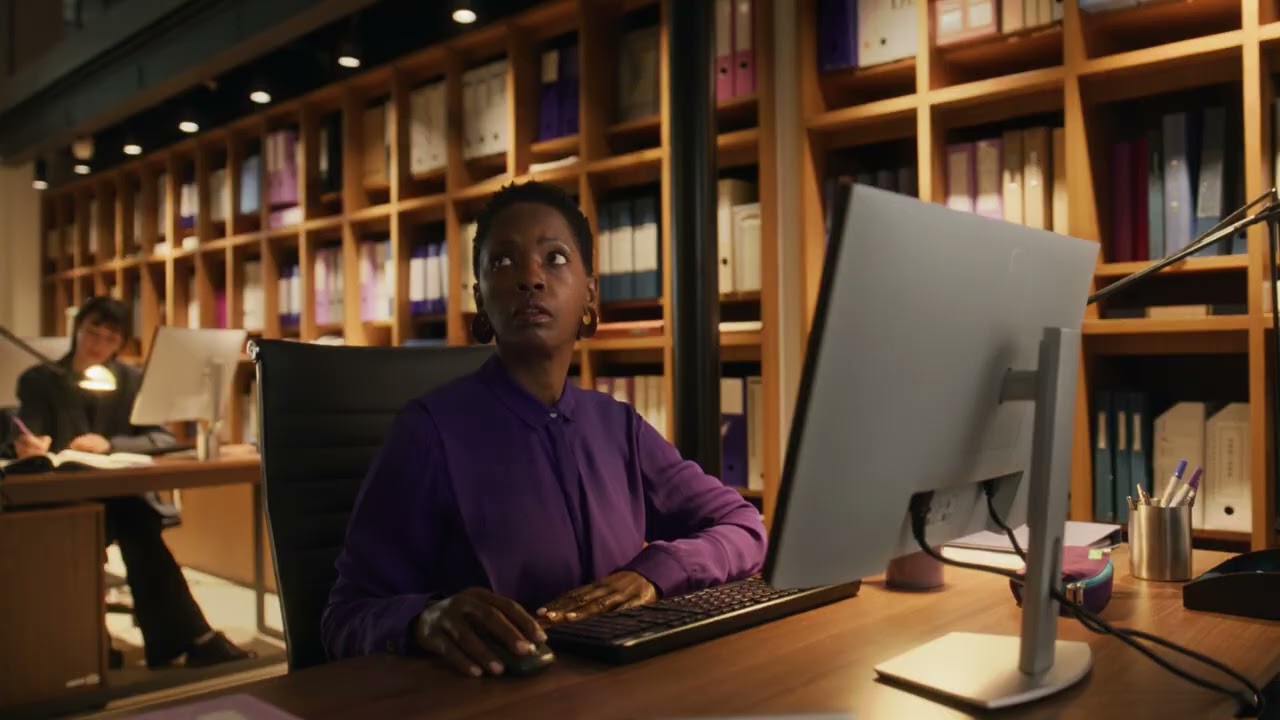 A woman in a purple shirt working at a desk in a modern office, using a desktop computer, with shelves of organized binders in the background.