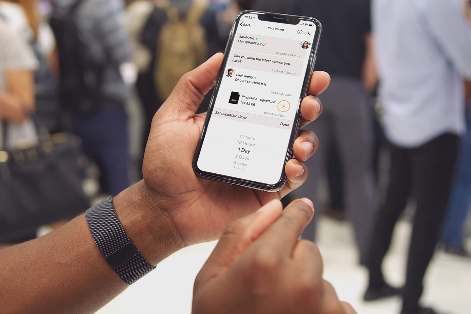 A person holding a smartphone displaying a messaging app interface, with chat messages and a file download option visible on the screen, in a crowded indoor setting.