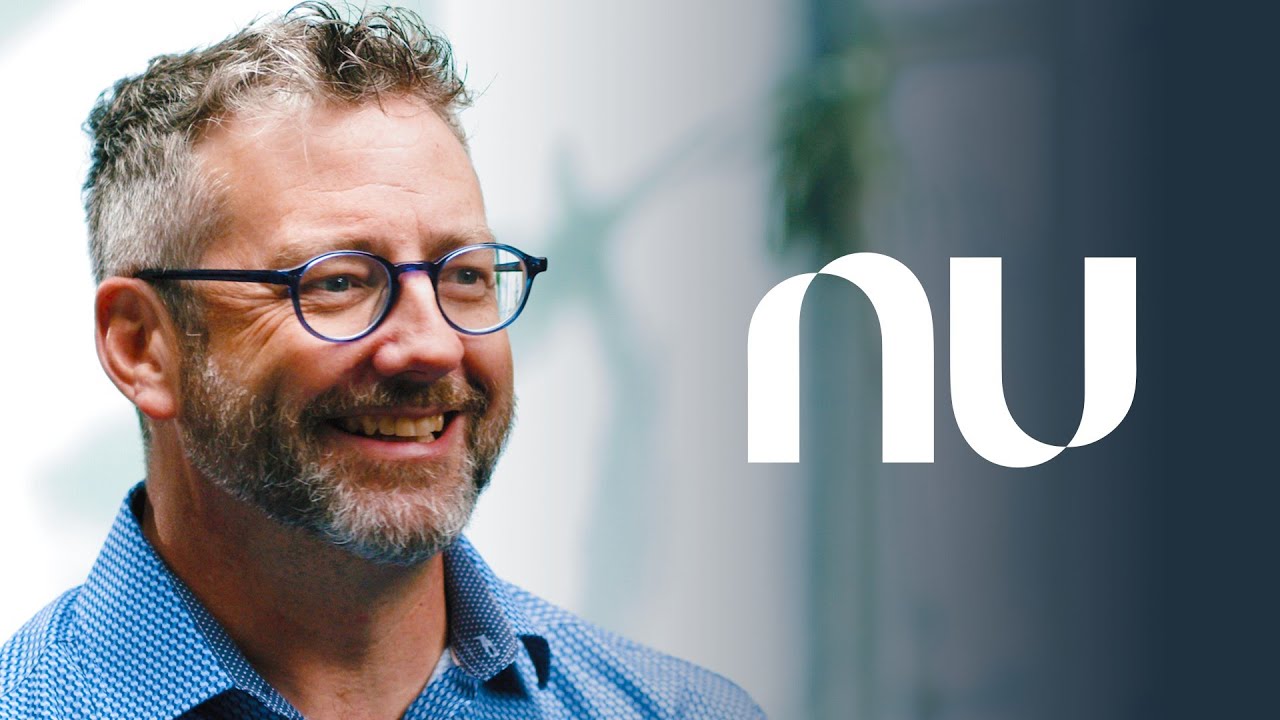 A smiling man with glasses and a beard wearing a blue shirt, with the NU logo displayed prominently to the right; designed as a YouTube thumbnail.
