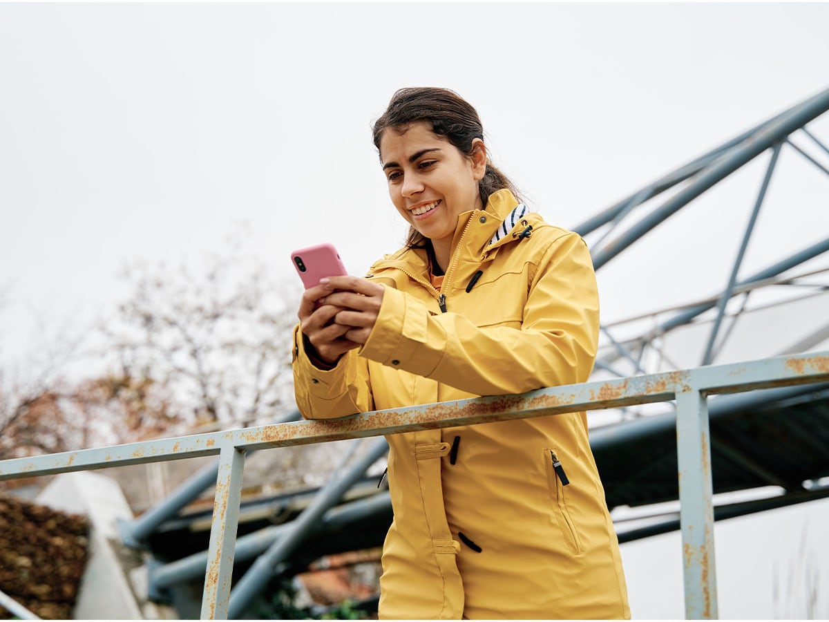 A woman wearing a yellow jacket stands outside, smiling while using a pink smartphone near a metal railing and modern structure.