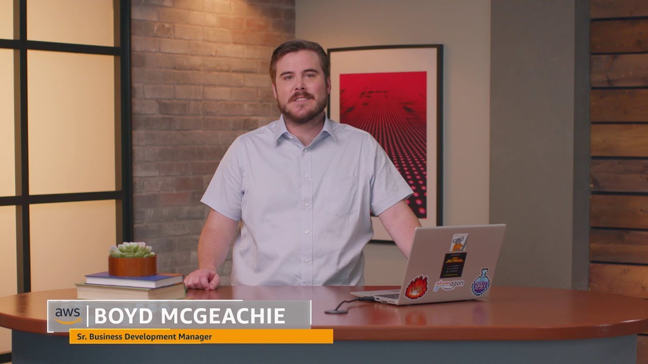 A person stands behind a desk with a laptop, books, and a plant, posing for a professional video or webcast. The setting appears to be an office or studio, with a modern background and wall art. This is a YouTube thumbnail for an AWS business development manager video.