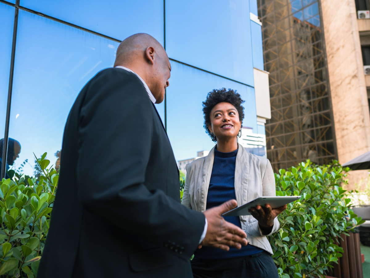 Two people standing outdoors near a modern glass building, one holding a tablet, surrounded by greenery.