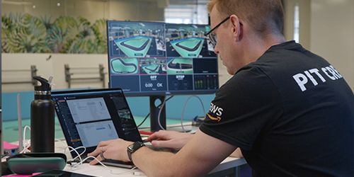 An engineer works at a desk, using a laptop and multiple monitors to design a bicycle using Autodesk software powered by AWS. The setup includes technical screens displaying bicycle models in a modern workspace.
