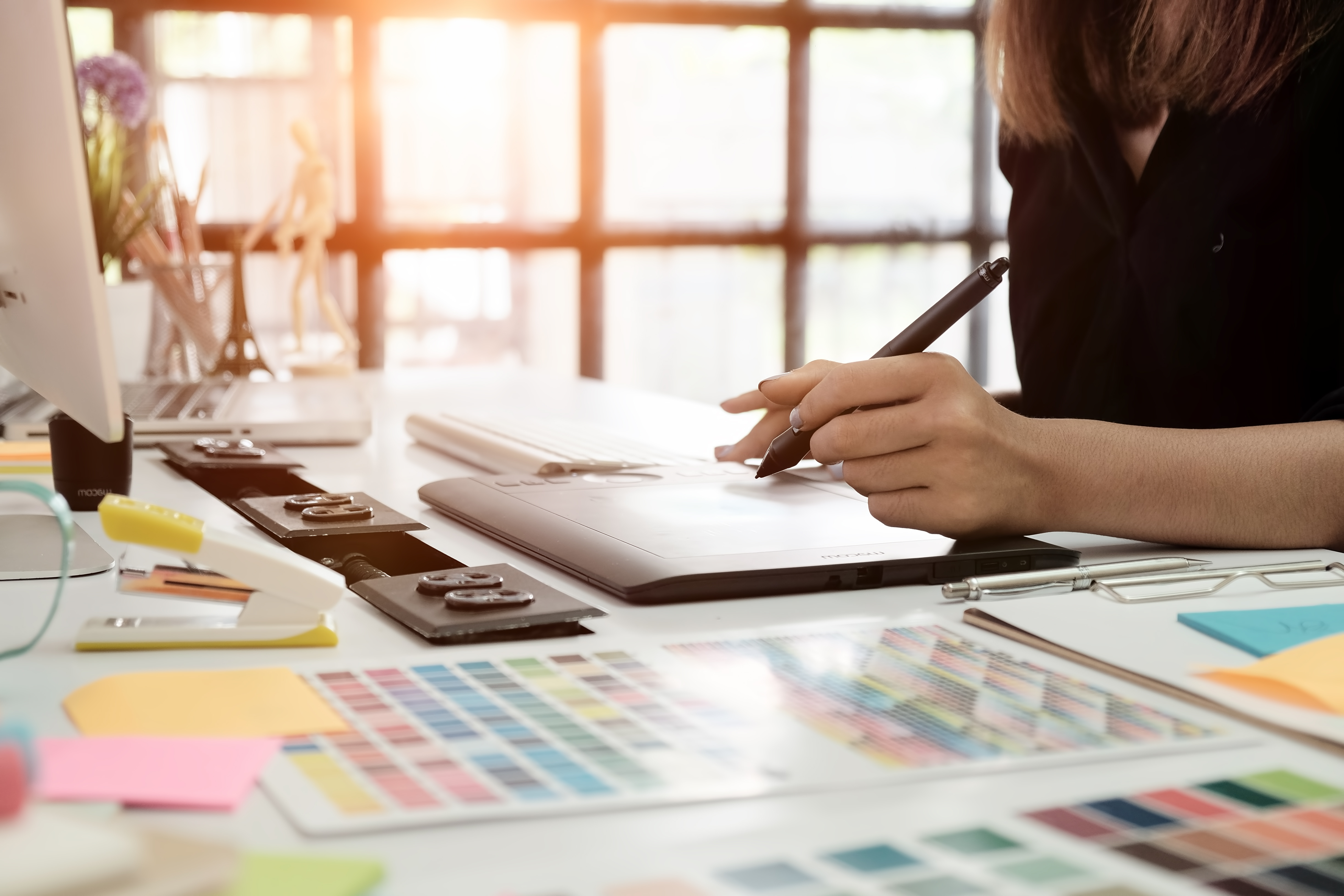 A graphic designer using a digital drawing tablet at a creative desk, surrounded by color swatches and design tools in a bright, modern workspace.