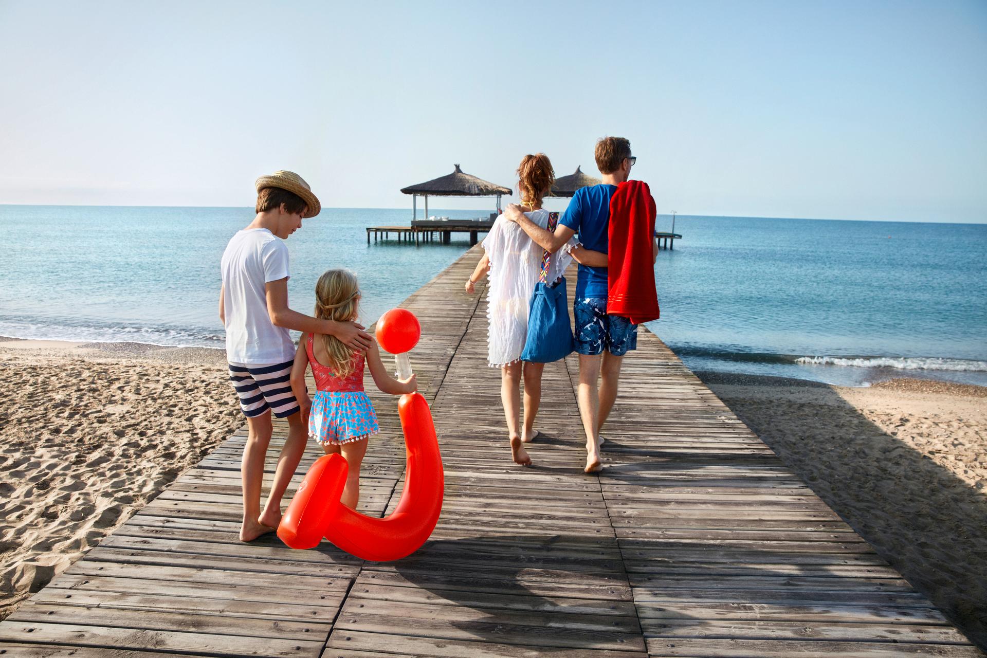 A family of four walking together on a wooden boardwalk leading to the ocean, with a sandy beach, clear blue sky, and two gazebos on the water. The children carry beach toys, evoking a sense of togetherness and summer vacation.