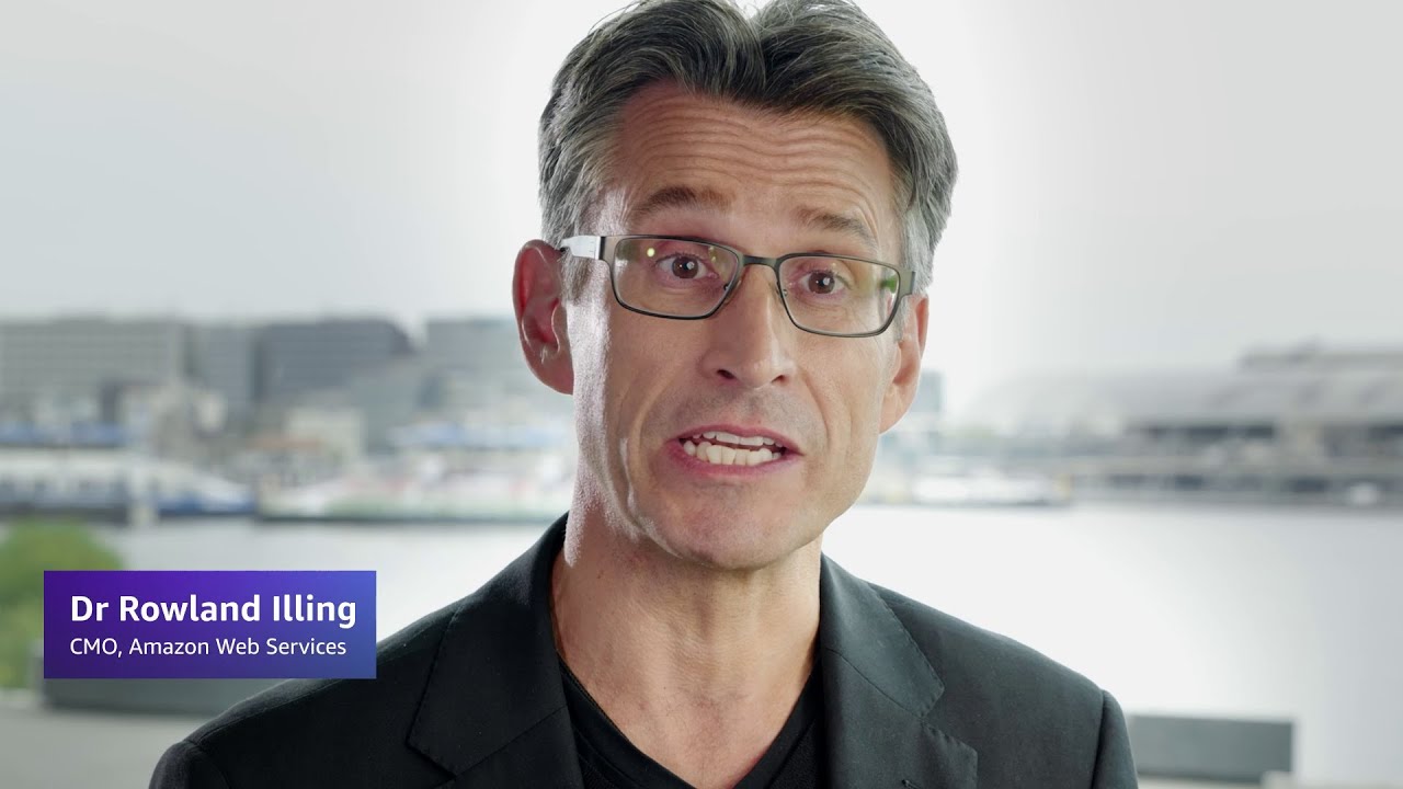 Dr. Rowland Illing, CMO of Amazon Web Services, with a blurred face, standing in front of a waterfront cityscape.
