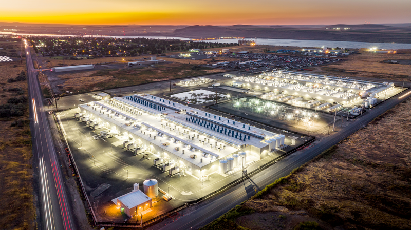 Aerial view of a brightly lit industrial facility at sunset, surrounded by open land and a distant river.