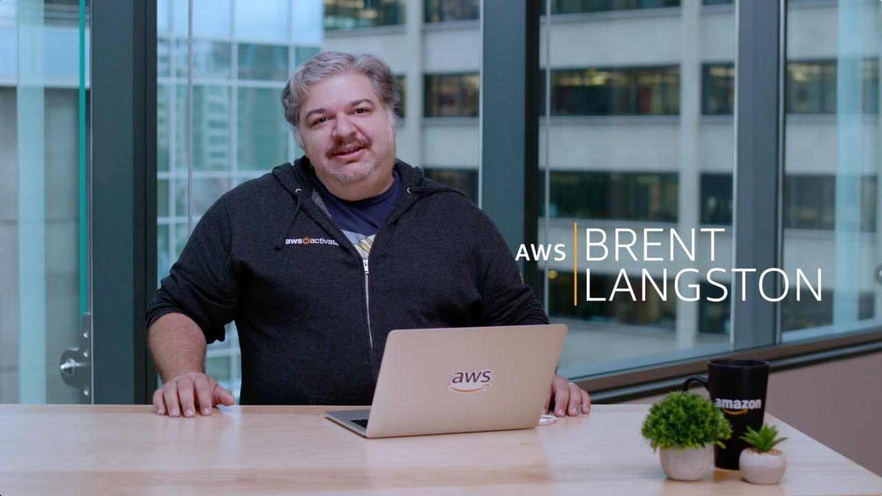 A person sits at a desk in an office setting with large windows, using a laptop with the AWS logo. There are plants and an Amazon mug on the desk. The text overlay reads 'AWS Brent Langston'.