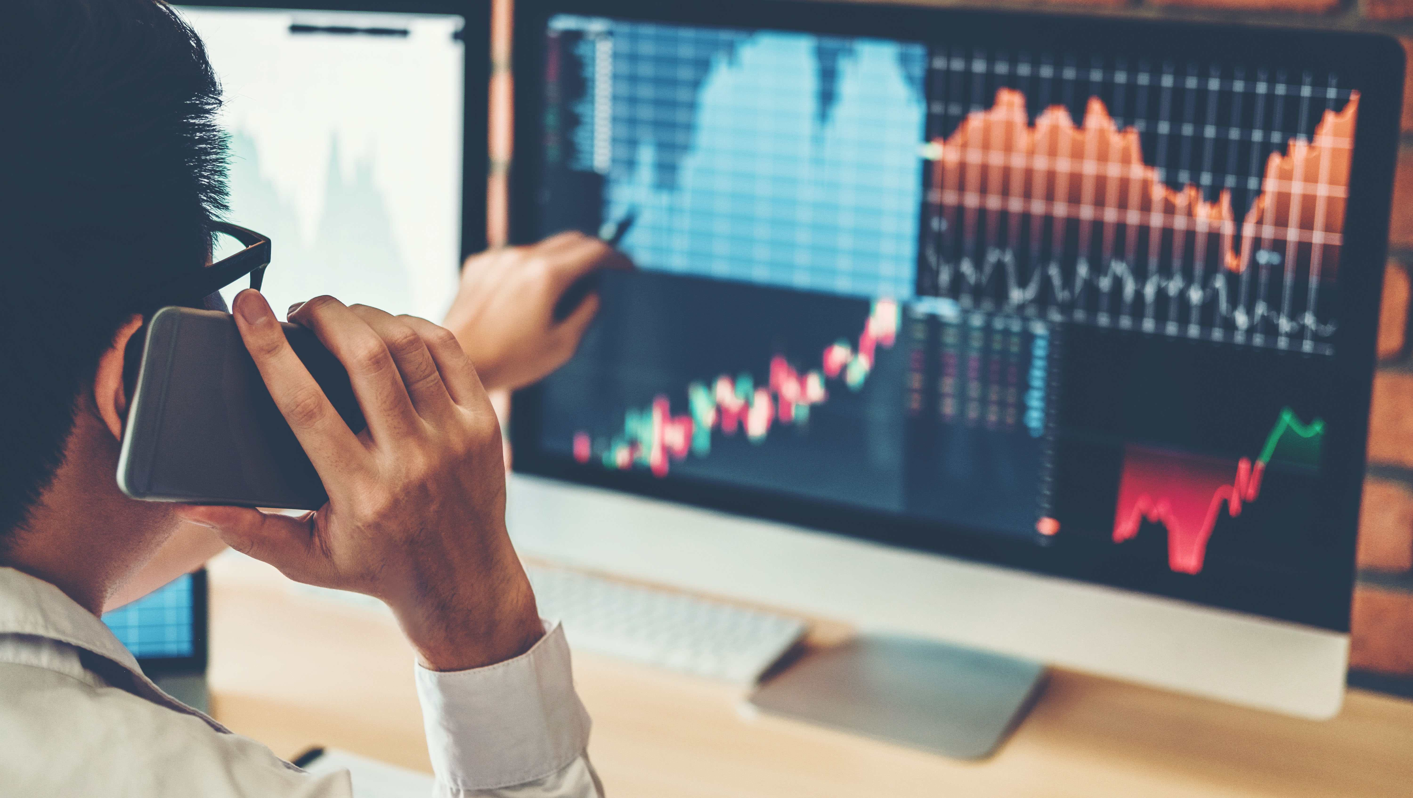 A person talks on a mobile phone while pointing at a monitor displaying stock market charts.