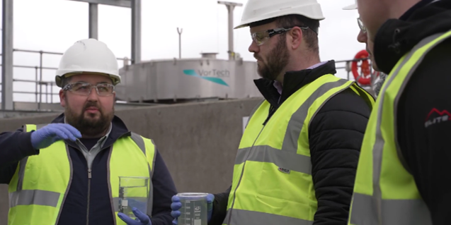 Engineers wearing hard hats, safety vests, and gloves inspect samples at a water treatment plant; 'VorTech' equipment branding is visible in the background.