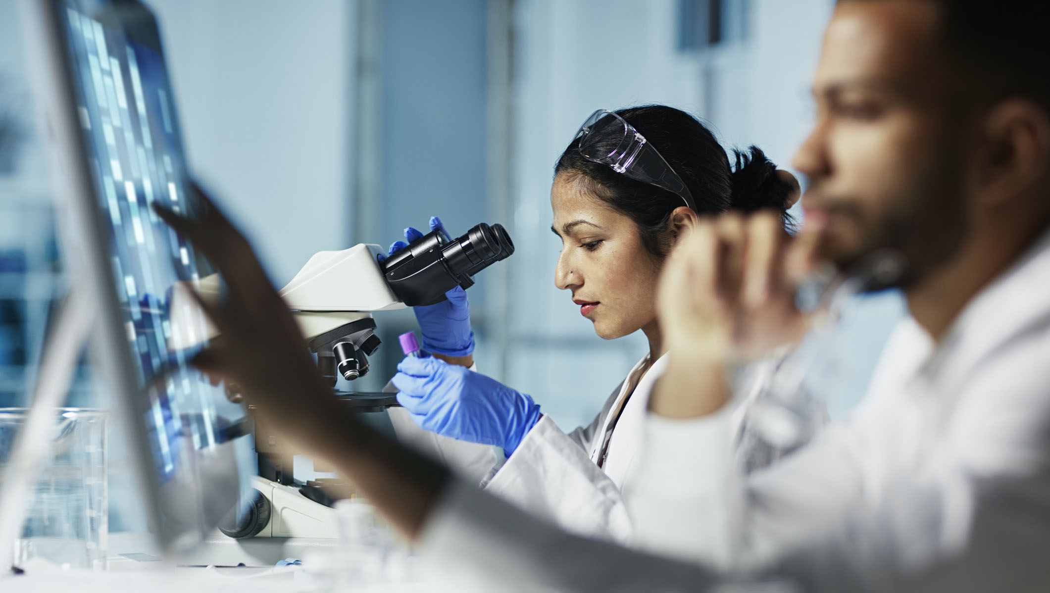Researchers in a laboratory observe data on a computer screen and use a microscope for scientific analysis. The scene highlights teamwork, technology, and scientific research in a modern lab environment.