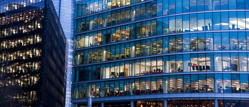 A modern office building with a glass exterior photographed in the evening. The image shows the illuminated interior of the offices through the glass windows, with multiple floors visible and the glow of office lights creating a vibrant urban atmosphere.