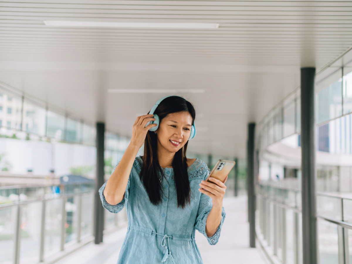 A woman wearing headphones is smiling while using her phone in a modern, glass-enclosed pedestrian walkway.