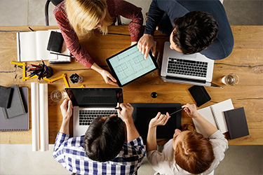 A group of people collaborating around a table with laptops and tablets, viewed from above.